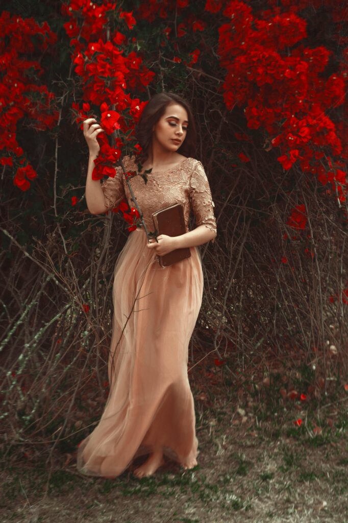pexels-photo-16564301-16564301 Woman in a flowing dress holding a book, surrounded by vibrant red flowers.